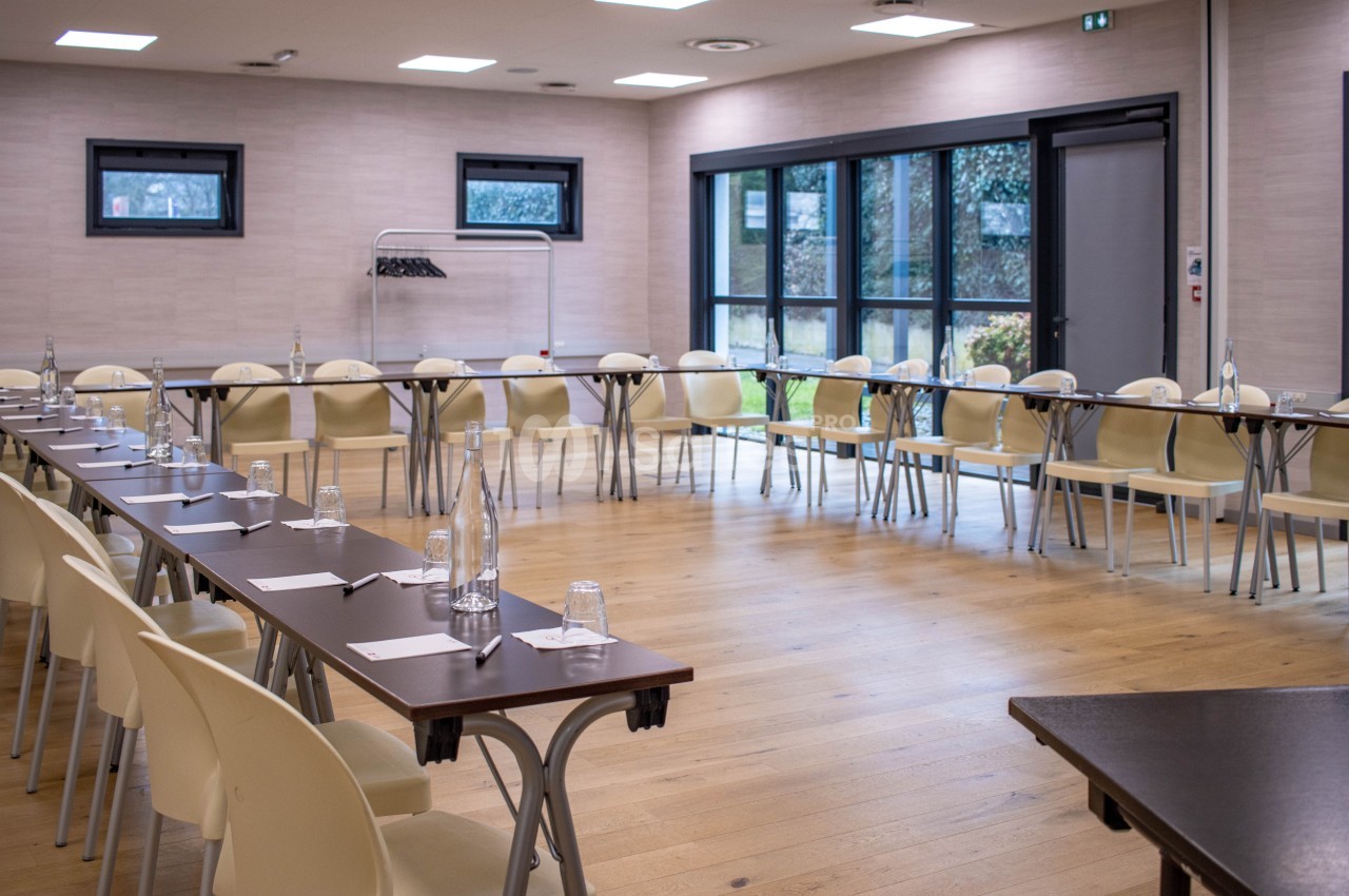 Salle de réunion lumineuse avec tables en U, chaises beige, bouteilles d'eau et fenêtres donnant sur un extérieur arboré.