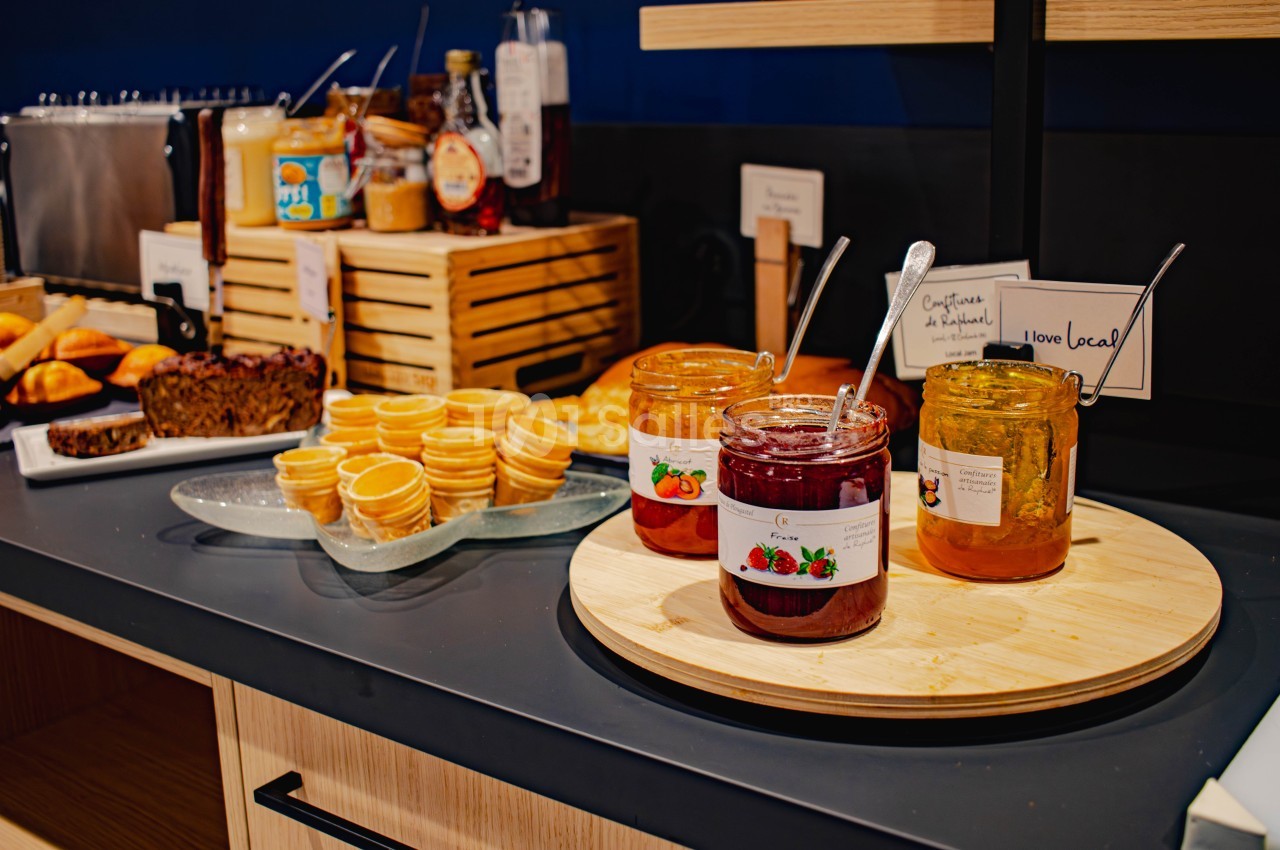Pots de confiture et miel sur un plateau en bois, accompagnés de petits gaufrettes et pâtisseries en arrière-plan.