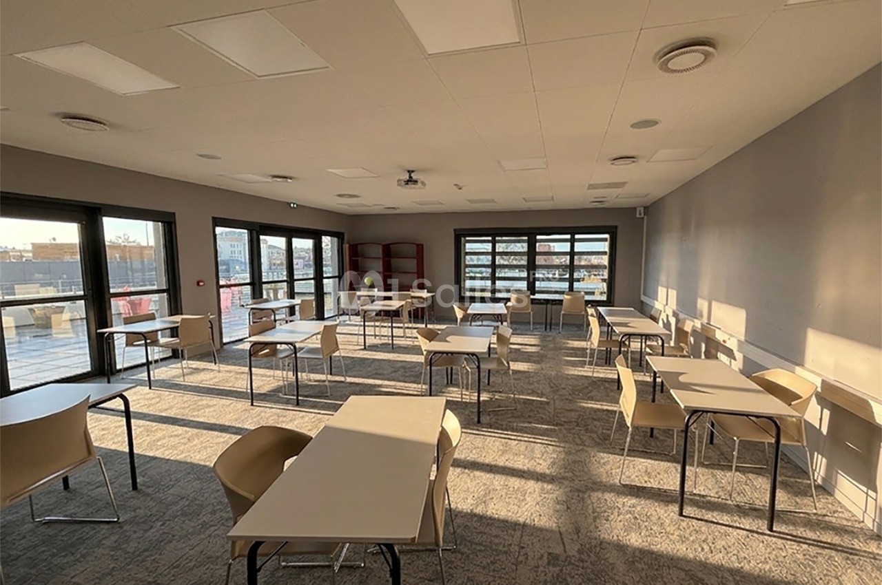 Salle lumineuse avec des tables et des chaises espacées, grandes fenêtres laissant entrer la lumière naturelle.