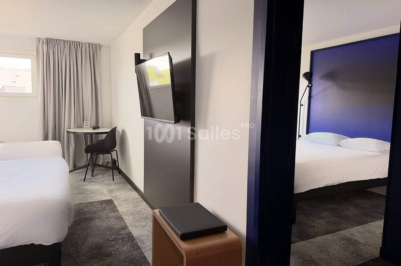 Chambre d'hôtel moderne avec deux lits simples, un bureau, une télévision murale et un aperçu d'une seconde pièce.