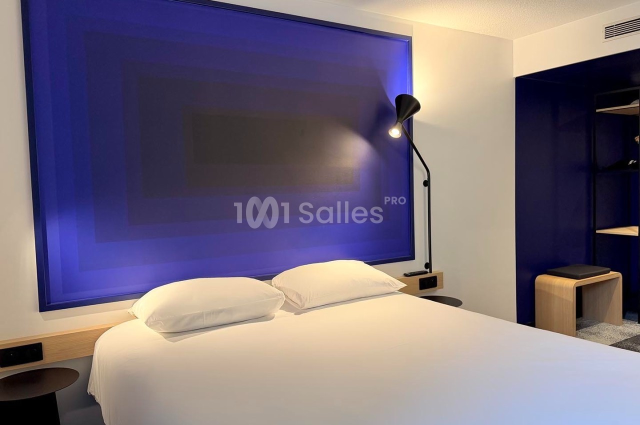 Lit double avec draps blancs, éclairé par une lampe noire, devant un panneau mural bleu à motifs géométriques.