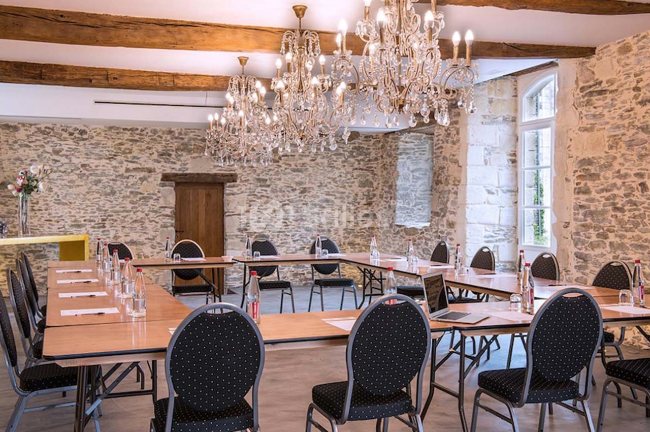 Salle de réunion lumineuse avec murs en pierre, tables disposées en U, chaises noires et lustres élégants.