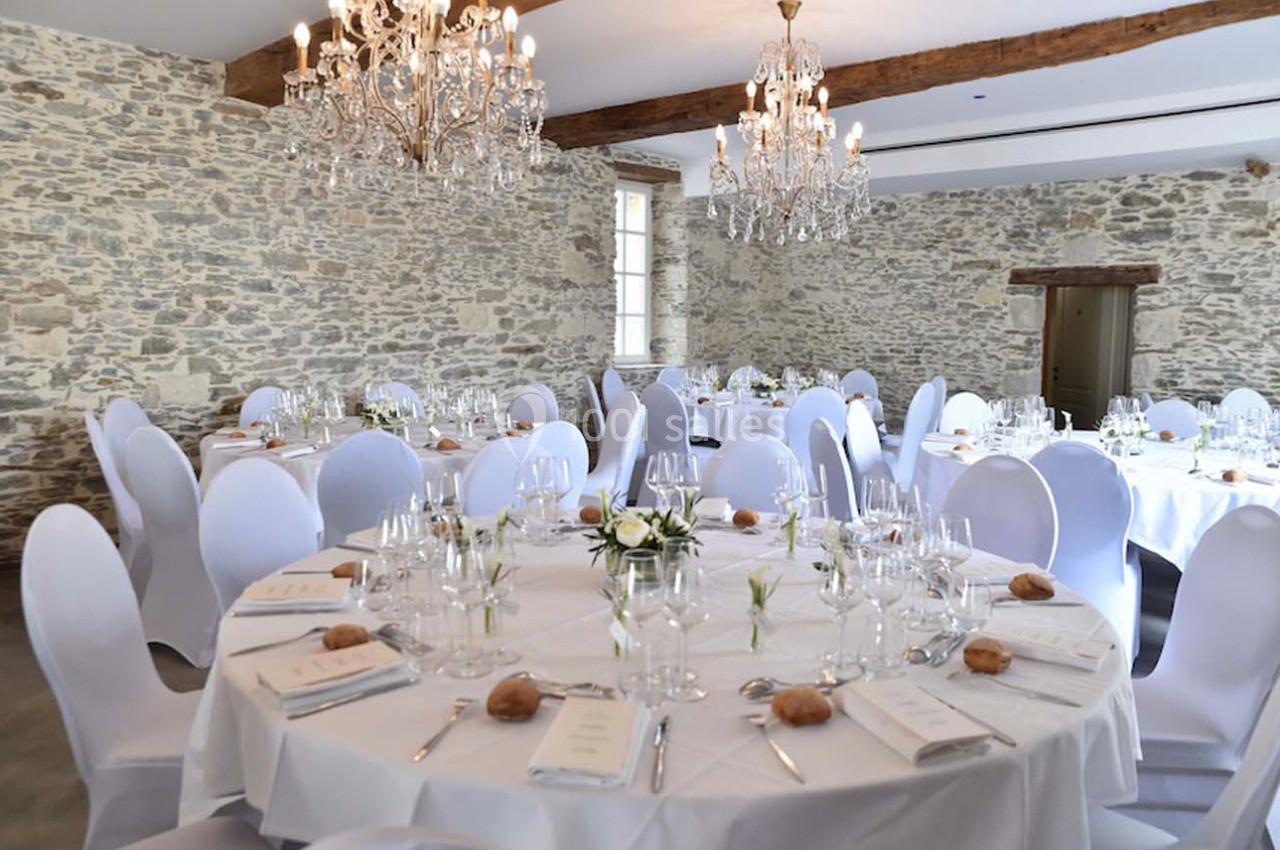 Salle de réception avec murs en pierre, tables rondes dressées avec nappes blanches, chandeliers et décoration élégante.
