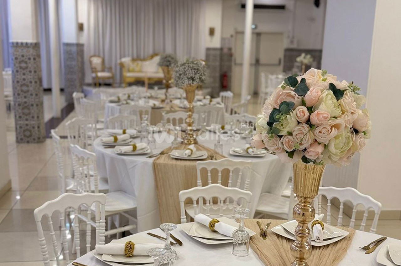 Salle de réception décorée avec des tables rondes, nappes blanches, chaises blanches et centres de table fleuris.