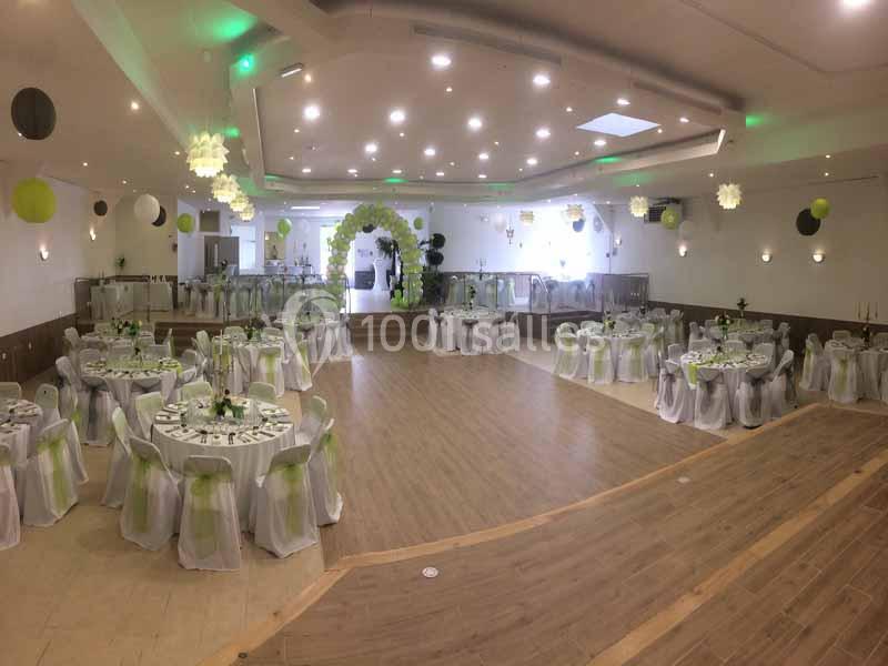 Salle de réception décorée avec des tables rondes, nappes blanches et accents verts, éclairage lumineux.