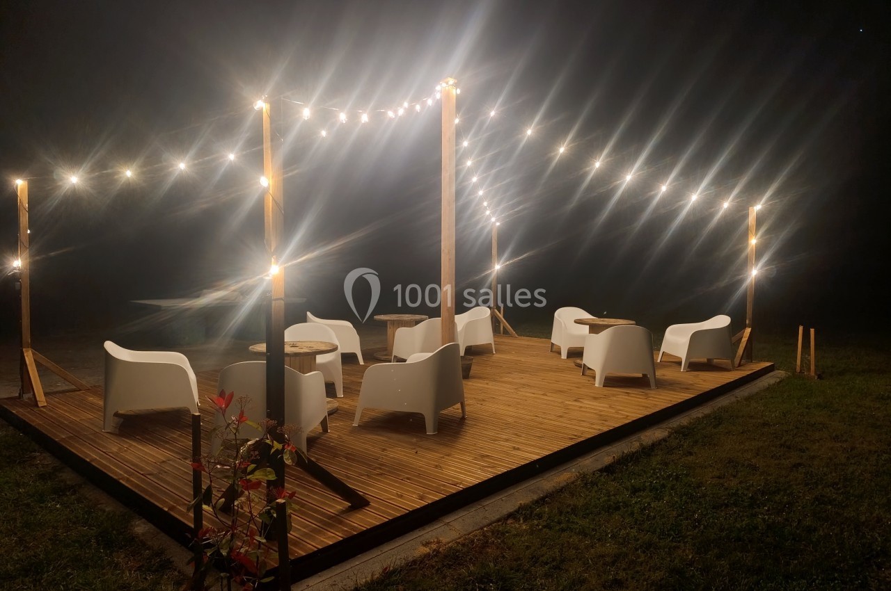 Terrasse en bois éclairée par des guirlandes lumineuses, avec des fauteuils et des tables disposés en extérieur.
