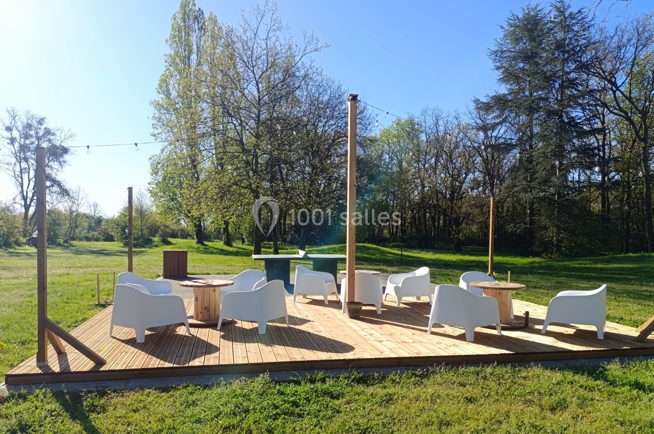 Terrasse en bois avec fauteuils blancs et tables basses, située dans un espace vert entouré d'arbres.