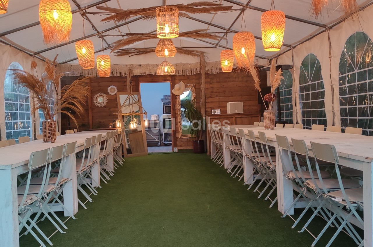 Intérieur d'une tente aménagée avec de longues tables blanches, des chaises pliantes et des lampes suspendues.