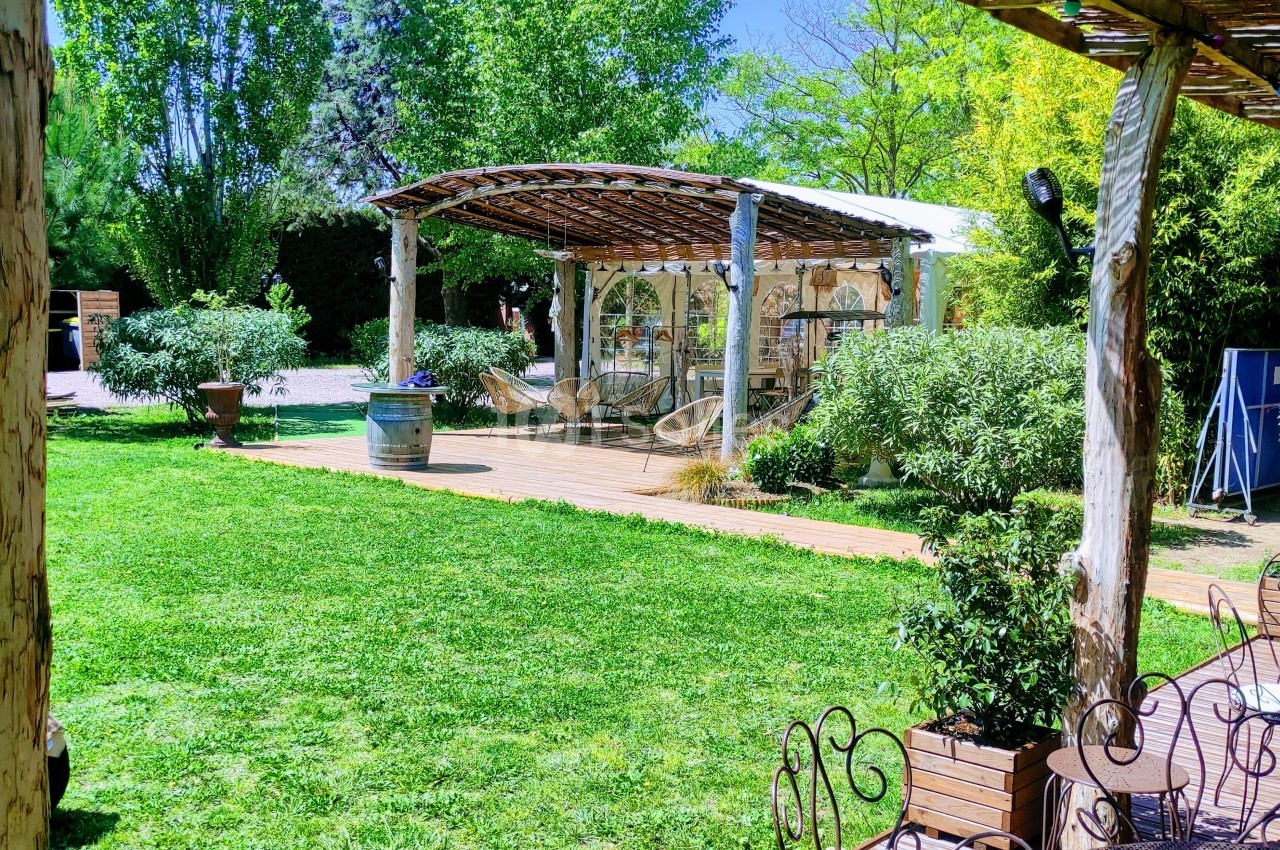 Espace extérieur avec pelouse, pergola en bois, chaises et végétation sous un ciel ensoleillé.