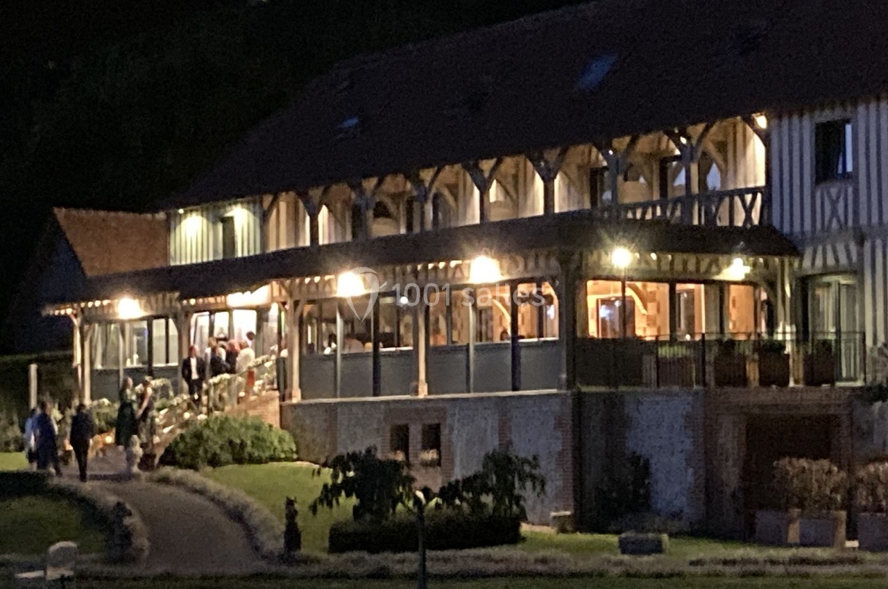 Bâtiment à colombages éclairé de nuit, avec terrasse animée et jardin aménagé au premier plan.
