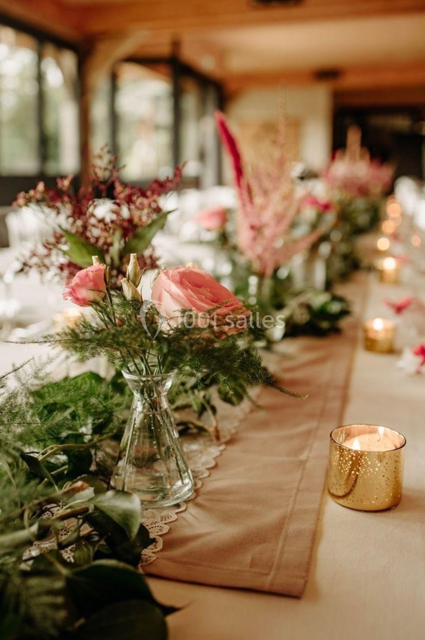 Centre de table avec fleurs roses et feuillages dans des vases en verre, bougies dorées allumées sur une nappe beige.