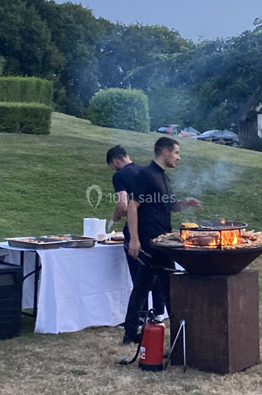 Deux hommes préparent un barbecue en plein air, avec une grande grille et une table dressée à proximité.