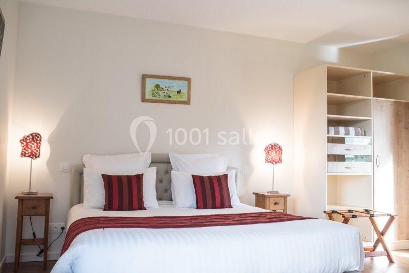 Lit double avec draps blancs, coussins rouges, tables de chevet avec lampes et armoire ouverte dans une chambre lumineuse.