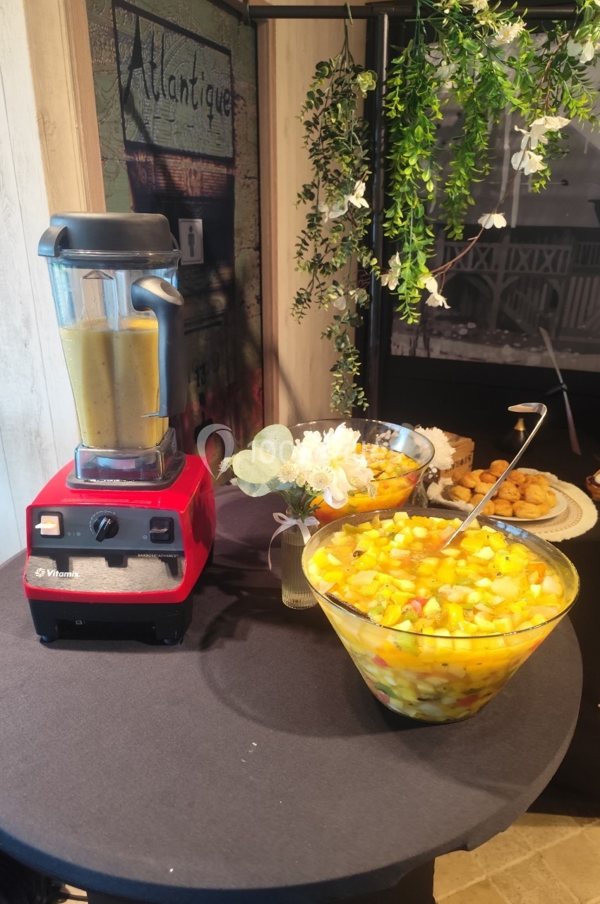 Un mixeur rouge à côté d'un grand saladier de salade de fruits sur une table décorée de fleurs.