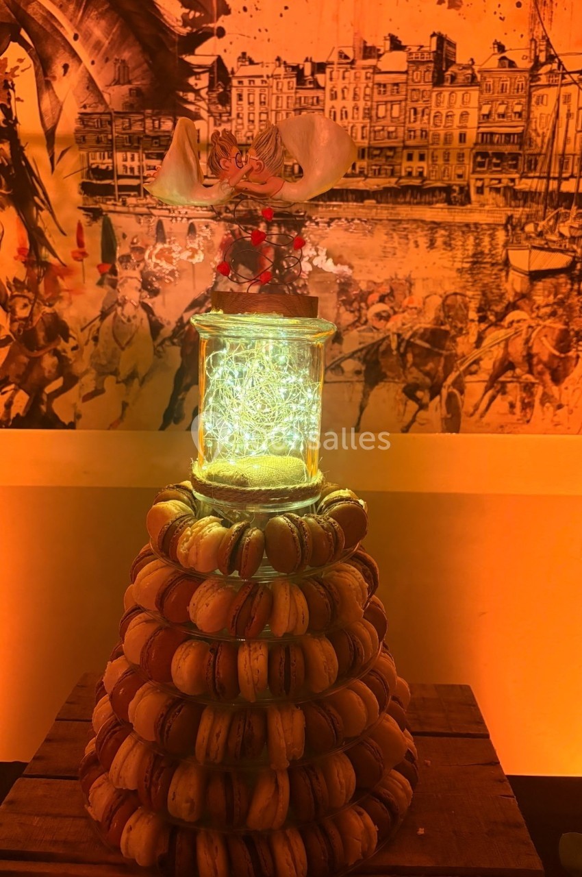 Tour de macarons disposés en pyramide, surmontée d'un bocal lumineux et d'une décoration florale, en arrière-plan un tableau…