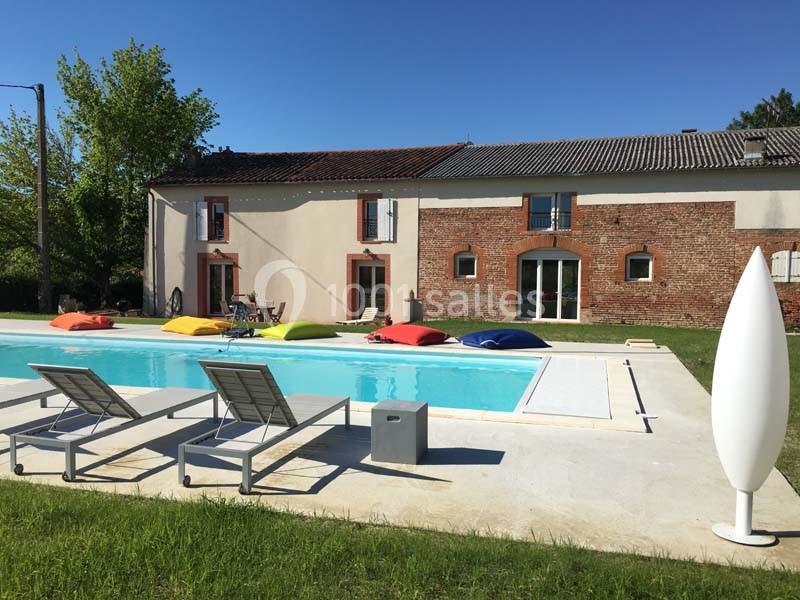 Maison en briques avec piscine extérieure, transats, coussins colorés et pelouse sous un ciel dégagé.