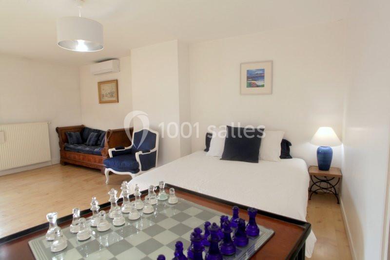 Chambre lumineuse avec lit double, canapé, fauteuil, échiquier au premier plan et décoration minimaliste.