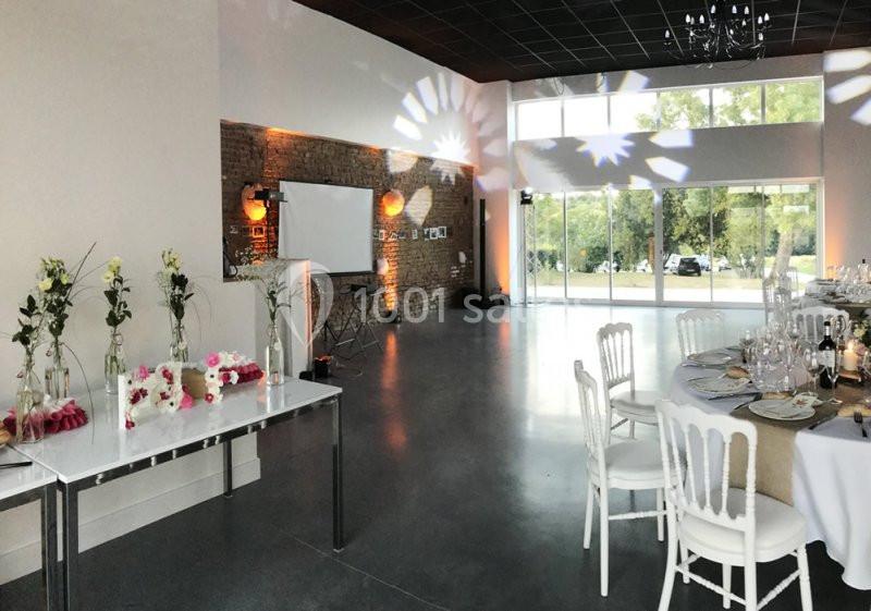 Salle de réception lumineuse avec tables dressées, décorations florales et grandes baies vitrées donnant sur l'extérieur.