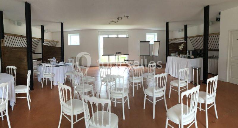 Salle lumineuse aménagée pour un événement, avec des chaises blanches, tables rondes et équipements de présentation.