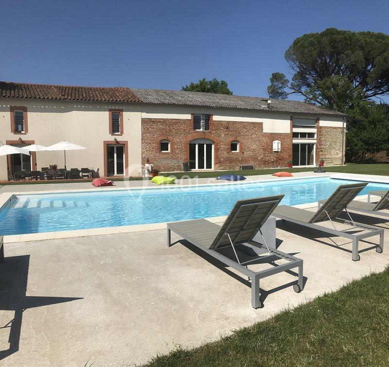 Piscine extérieure avec transats devant une grande maison en briques et crépi, entourée de verdure.