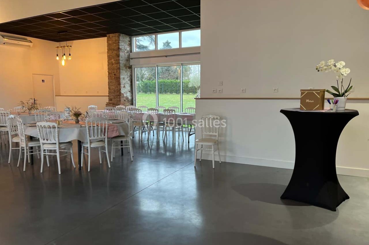 Location salle Sainte-Foy-d'Aigrefeuille (Haute-Garonne) - Les Ecuries de Ronsac #6 Salle de réception lumineuse avec tables dressées, chaises blanches, grandes baies vitrées et un pupitre décoré.