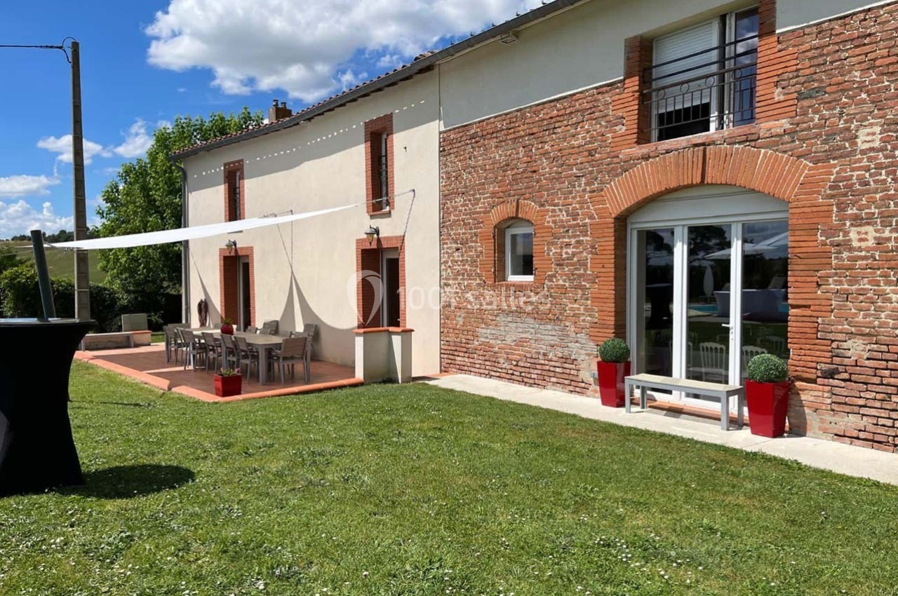 Location salle Sainte-Foy-d'Aigrefeuille (Haute-Garonne) - Les Ecuries de Ronsac #2 Façade d'une maison en briques et crépi avec terrasse aménagée, pelouse et ciel dégagé en arrière-plan.