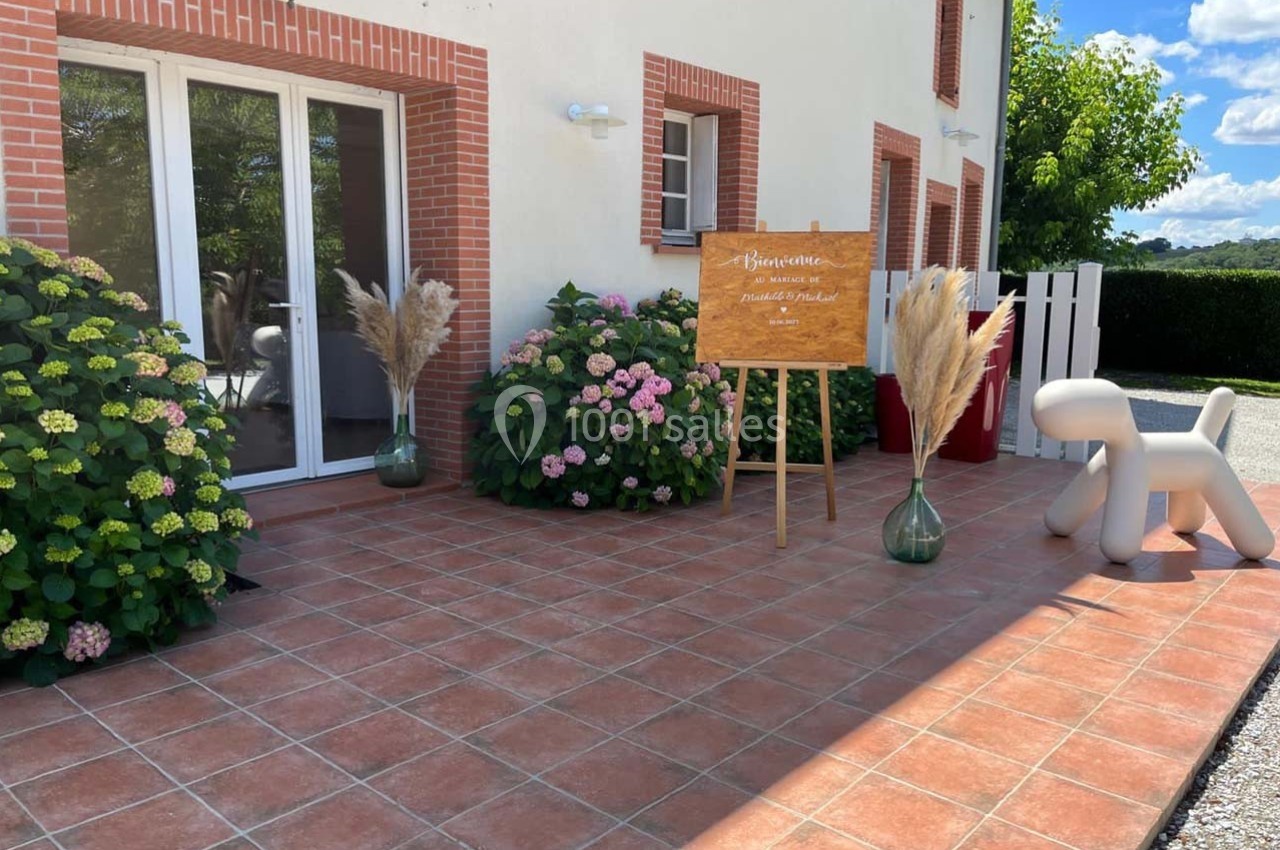 Location salle Sainte-Foy-d'Aigrefeuille (Haute-Garonne) - Les Ecuries de Ronsac #1 Terrasse carrelée avec plantes, panneau de bienvenue sur chevalet, vases décoratifs et siège en forme de chien.