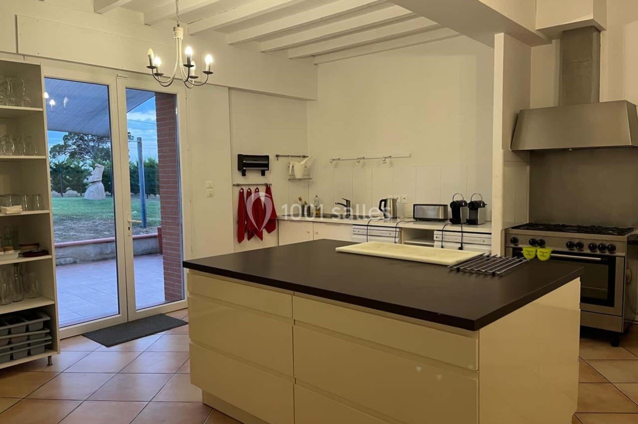 Location salle Sainte-Foy-d'Aigrefeuille (Haute-Garonne) - Les Ecuries de Ronsac #10 Cuisine moderne avec îlot central, plan de travail sombre, électroménagers en inox et vue sur une terrasse extérieure.