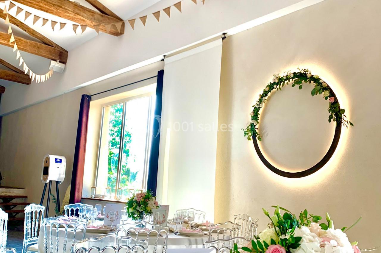 Salle de réception lumineuse décorée pour un mariage, avec tables dressées, guirlandes et couronne florale murale.