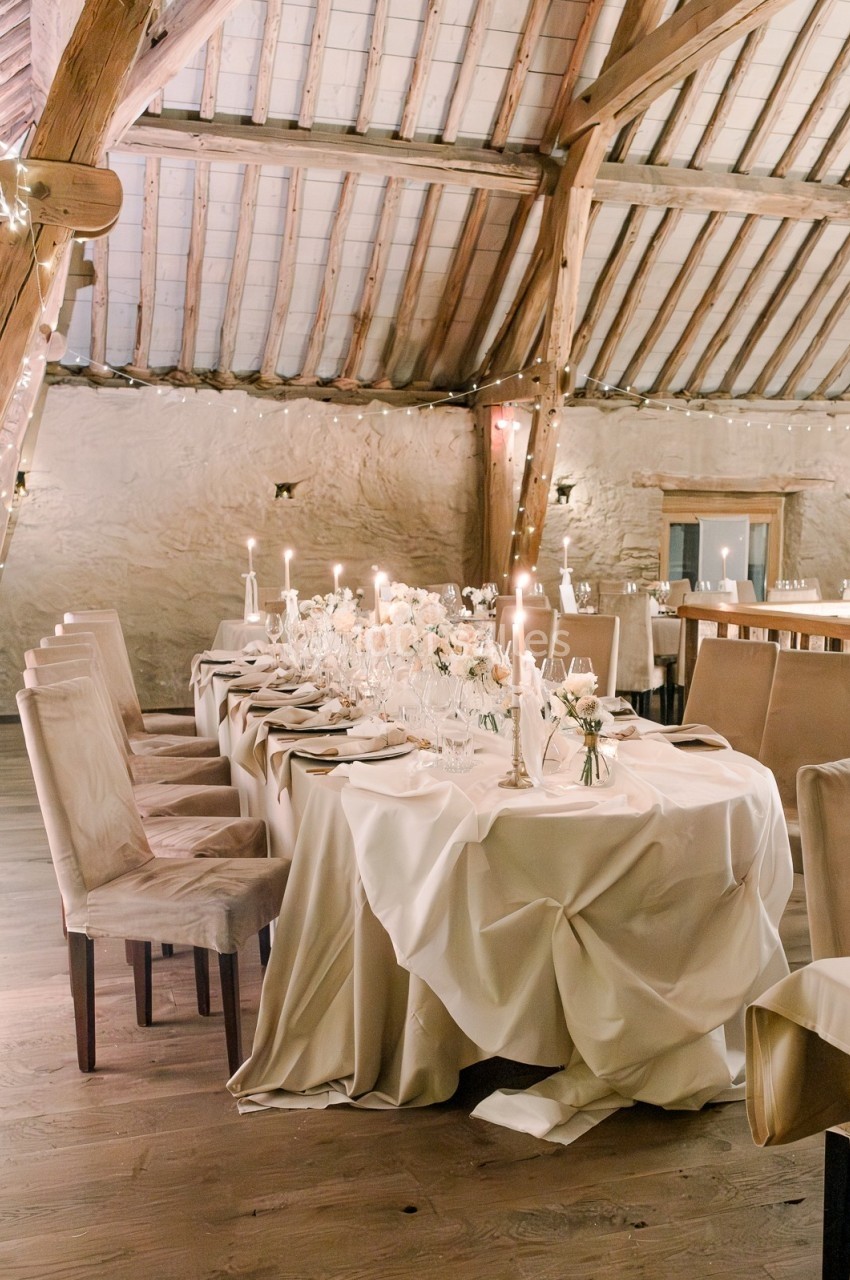 Table élégamment dressée avec nappes blanches, bougies et décorations florales, dans une salle en bois au plafond voûté.