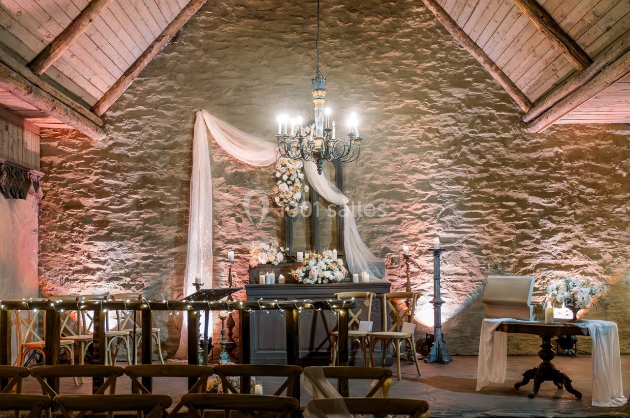 Salle en pierre avec un autel décoré de fleurs et de voilages, éclairée par un lustre et des lumières tamisées.
