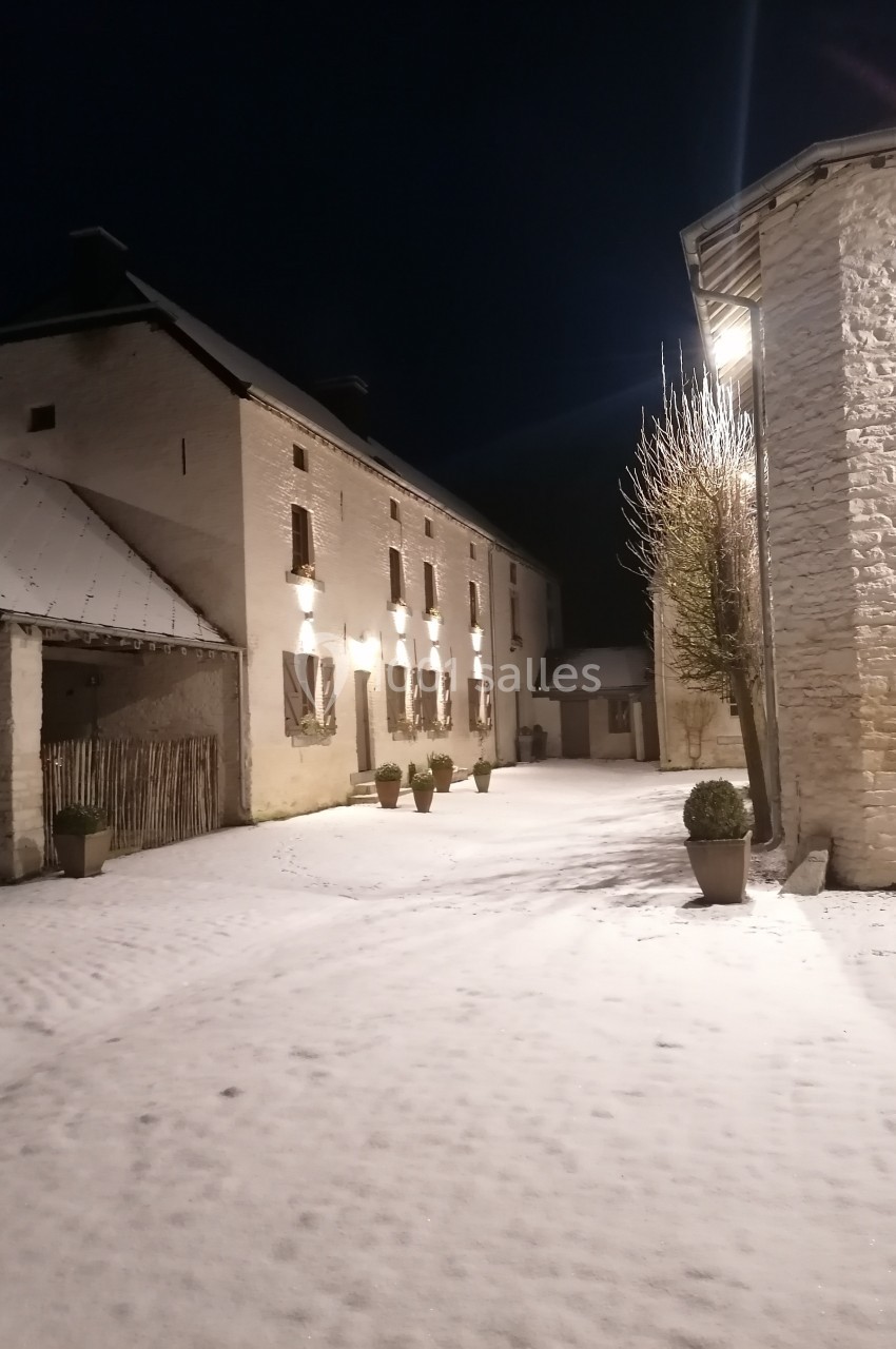 Cour enneigée de nuit entourée de bâtiments en pierre éclairés par des lampes murales.