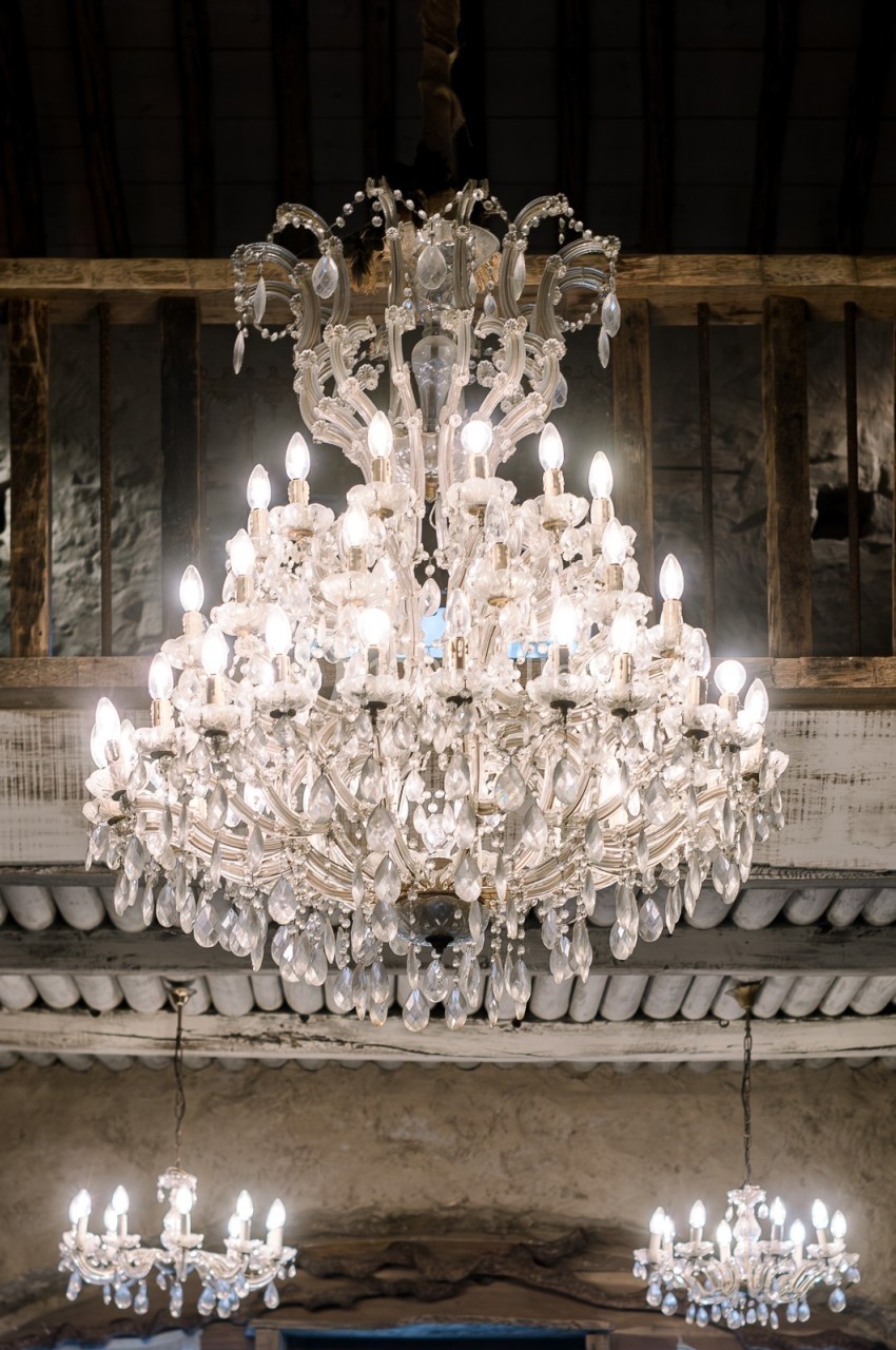 Lustre en cristal éclairé suspendu dans une pièce au plafond en bois et poutres apparentes.