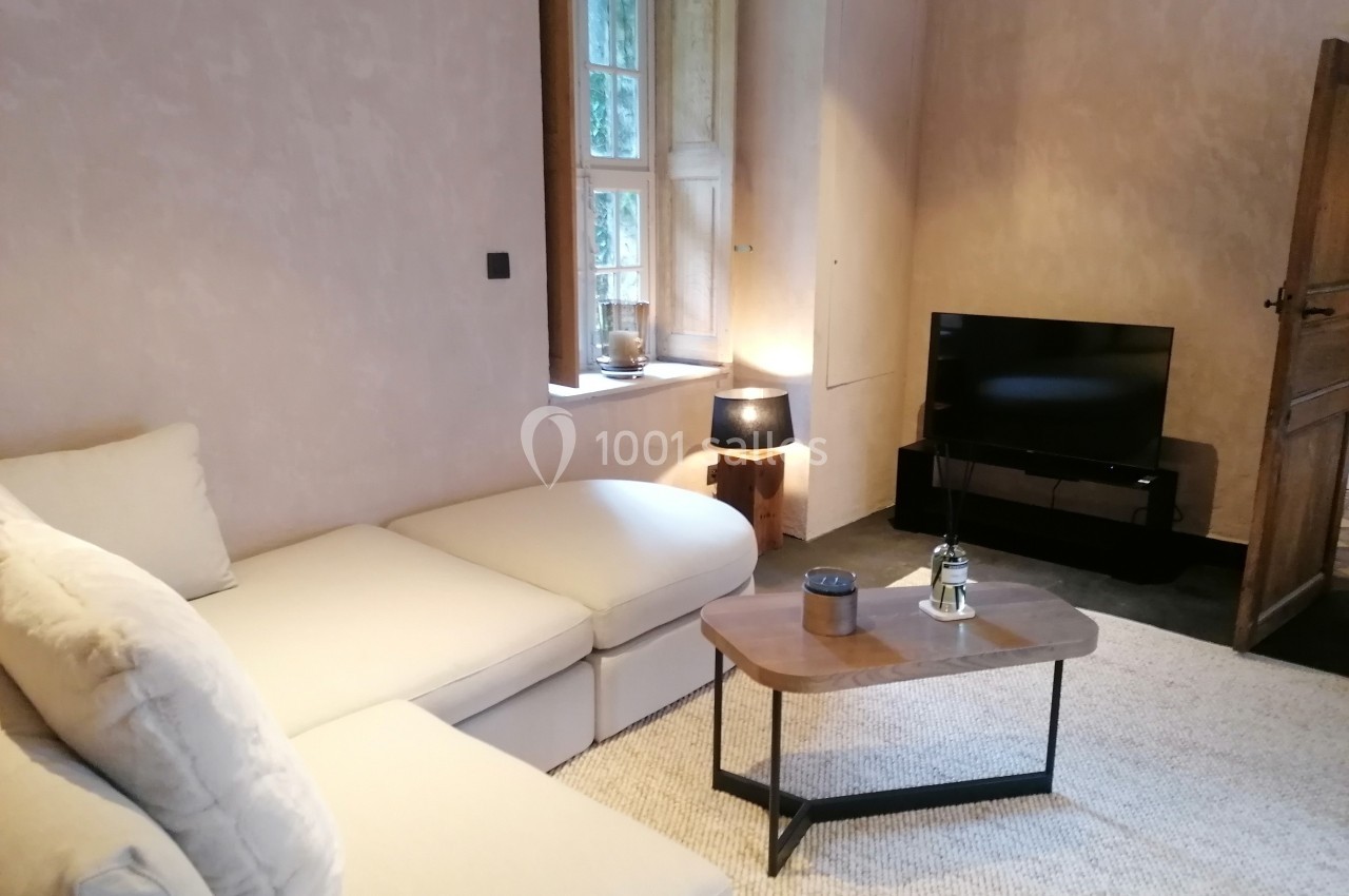 Salon avec canapé d'angle beige, table basse en bois, lampe, télévision et fenêtre laissant entrer la lumière naturelle.