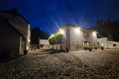 Cour pavée d'un bâtiment en pierre éclairé par des lumières extérieures, entouré d'arbres et de bâtiments annexes, de nuit.