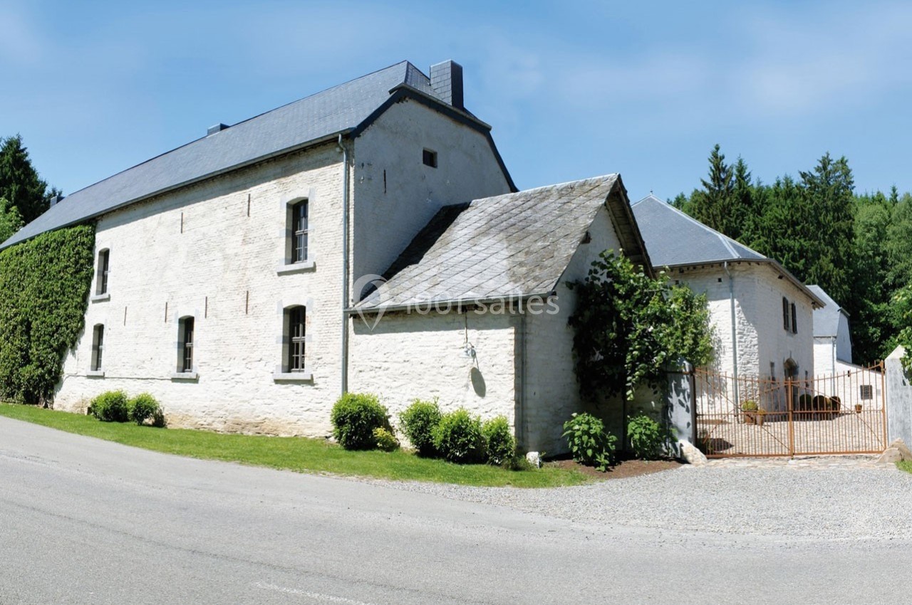 Bâtiment en pierre blanche avec toit en ardoise, entouré de verdure, situé près d'une route asphaltée.