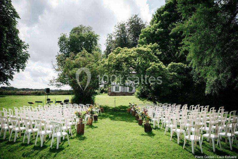 Location salle Saint-Romain-de-Colbosc (Seine-Maritime) - Château de Gromesnil #12 Chaises blanches disposées en rangées sur une pelouse, formant une allée menant à une petite maison à colombages.
