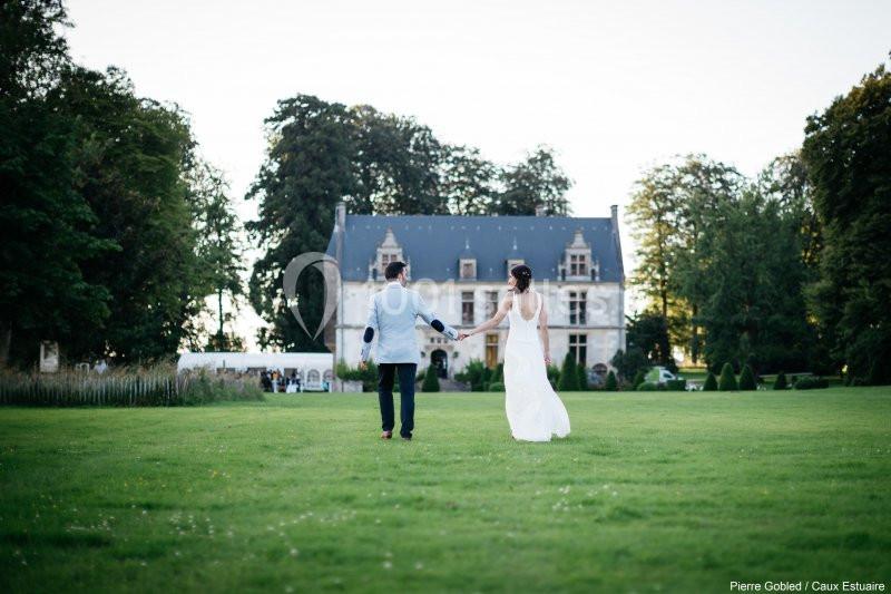 Location salle Saint-Romain-de-Colbosc (Seine-Maritime) - Château de Gromesnil #9 Un couple marche main dans la main sur une pelouse, avec un château en arrière-plan entouré d'arbres.
