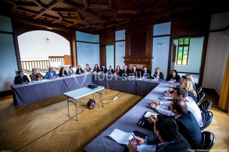 Réunion de plusieurs personnes assises autour d'une grande table en U dans une salle lumineuse avec parquet et boiseries.