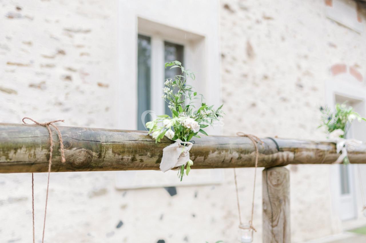 Arrangement floral suspendu sur une structure en bois devant un mur en pierre avec une fenêtre en arrière-plan.