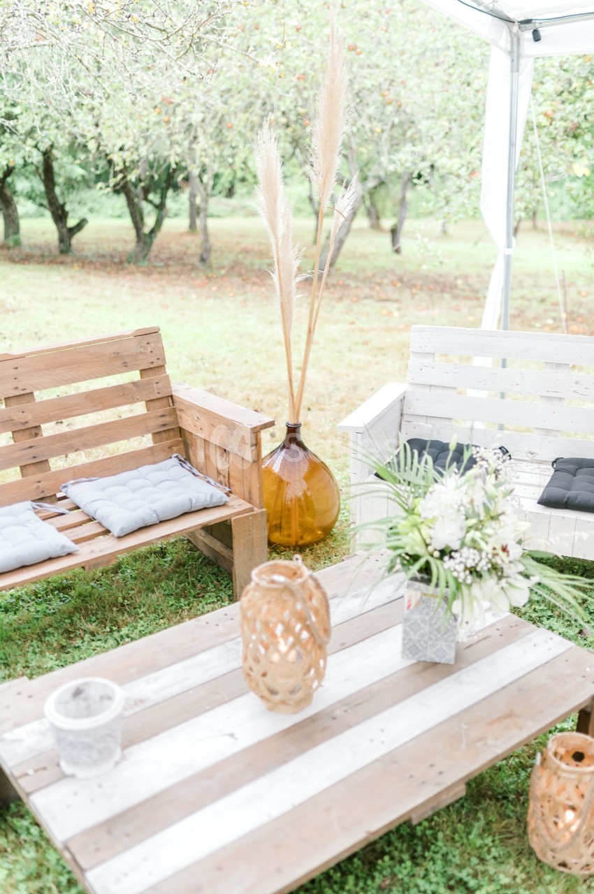 Salon de jardin en bois avec coussins, table basse décorée de fleurs et lanternes, installé dans un verger.