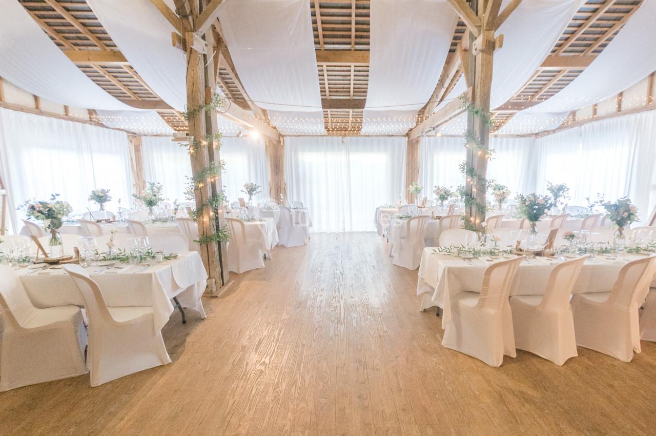 Salle de réception lumineuse décorée pour un événement, avec tables dressées et chaises blanches.