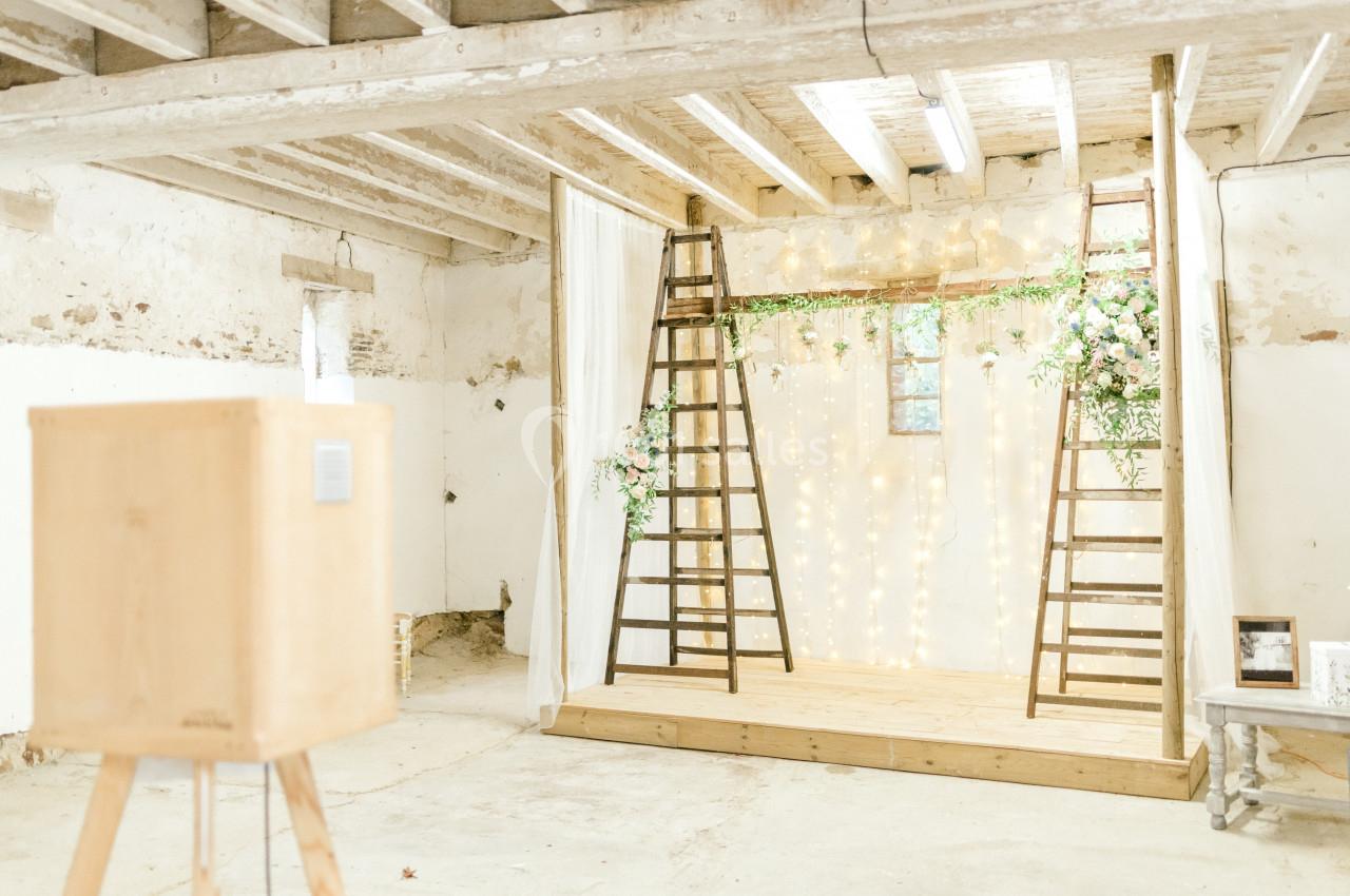 Scène de mariage rustique avec guirlandes lumineuses, échelle décorative et fleurs dans une grange lumineuse.