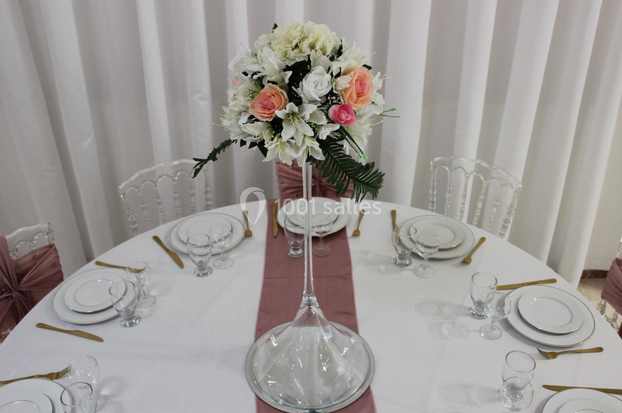 Centre de table floral avec roses et feuillage, posé sur une table ronde dressée avec vaisselle blanche et couverts dorés.