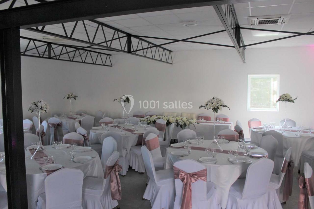 Salle de réception décorée avec des tables rondes, nappes blanches, chaises ornées de rubans roses et arrangements floraux.
