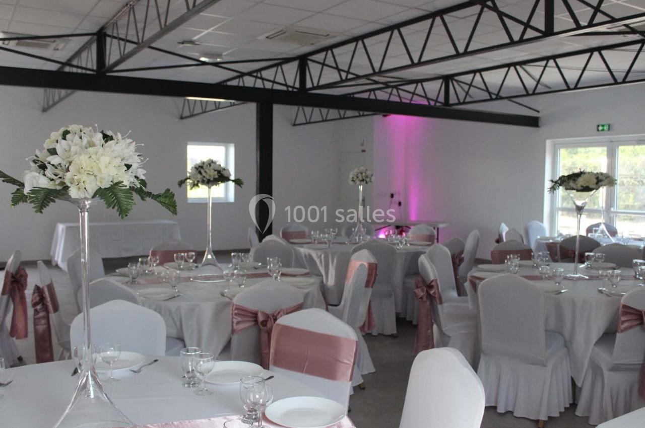 Salle de réception décorée avec des tables rondes, nappes blanches, chaises ornées de nœuds roses et centres de table…