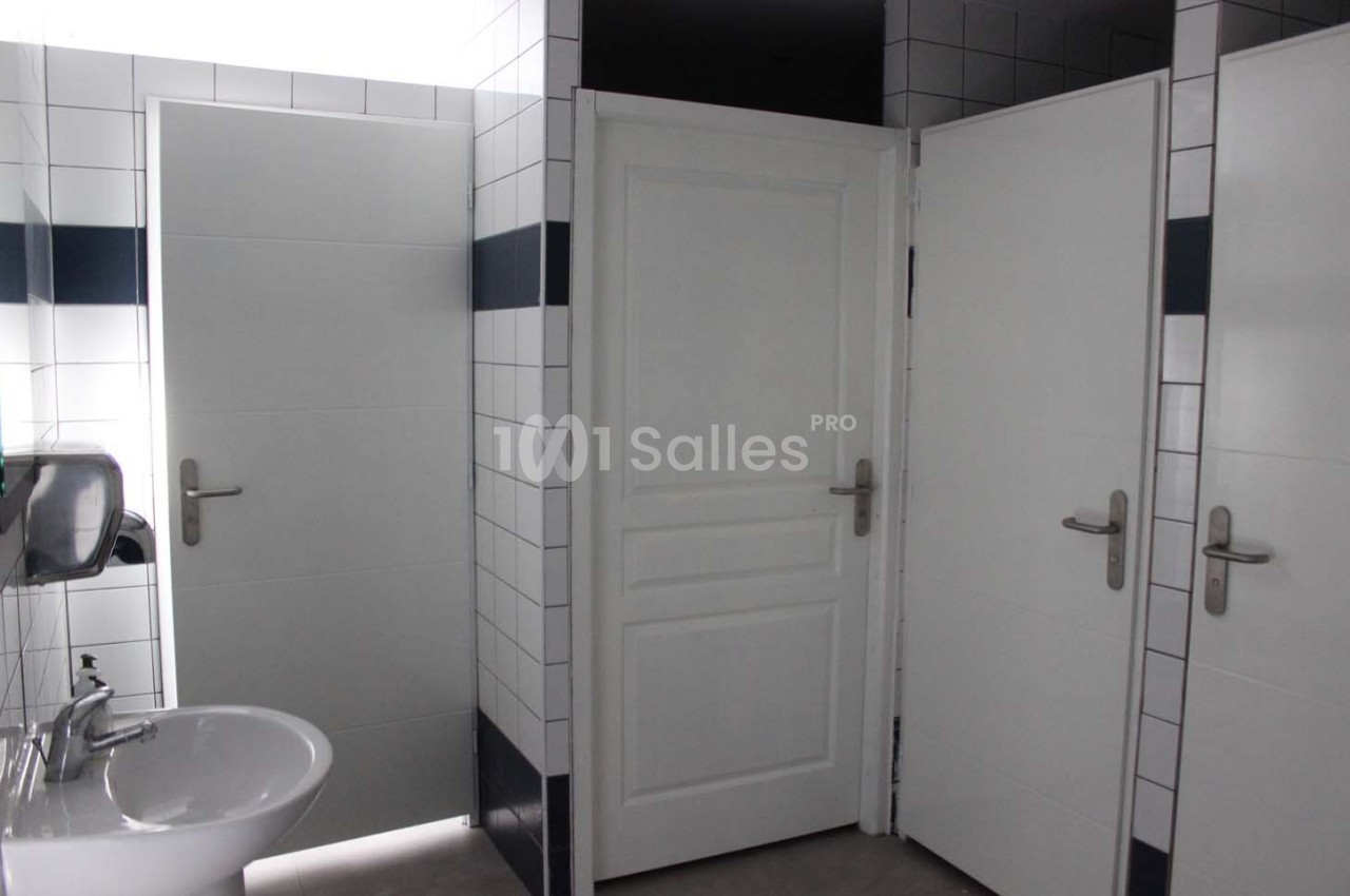 Portes de cabines de toilettes blanches dans un espace carrelé avec un lavabo et un distributeur de savon.