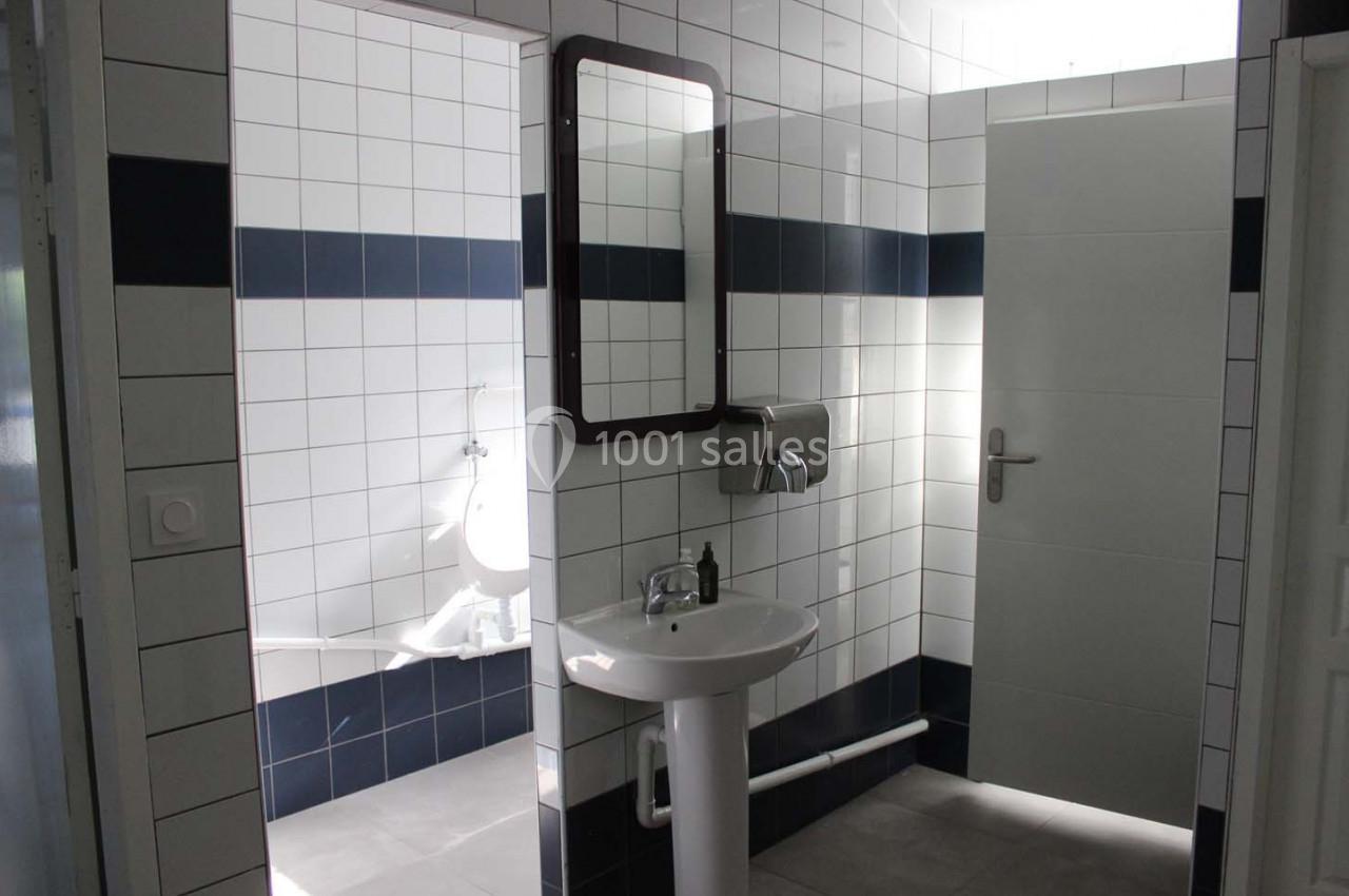 Lavabo avec miroir dans des toilettes carrelées blanches et bleues, avec urinoir visible en arrière-plan.