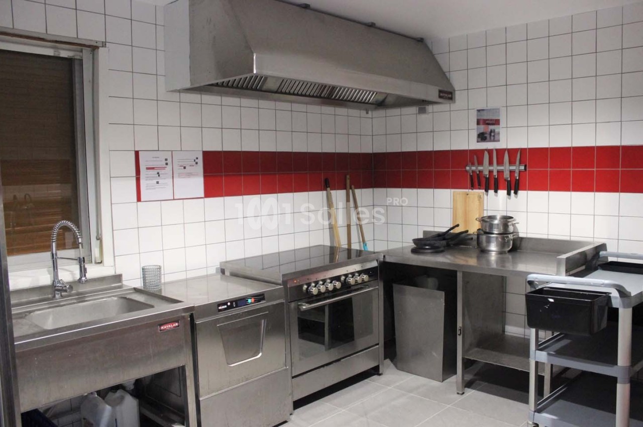 Cuisine professionnelle équipée avec évier, four, plaques de cuisson, hotte, plan de travail en inox et ustensiles accrochés.