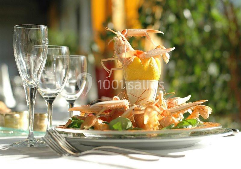 Assiette de langoustines dressées autour d'un citron sur un lit de verdure, avec des verres en arrière-plan.